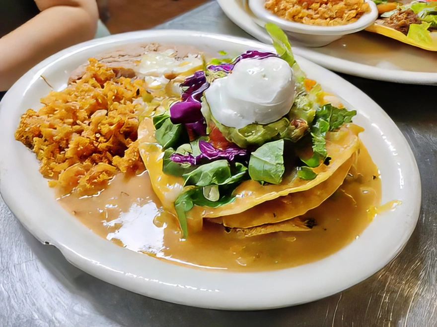 Santa Fe Three corn tortillas fried crisp and layered with ground sirloin, shredded chicken, or machaca. Smothered in chile verde or enchildad sauce. Topped with cheddar and Monterey jack cheese, lettuce, tomato, sour cream, and guacamole. Served with spanish rice or fideo and refried beans.